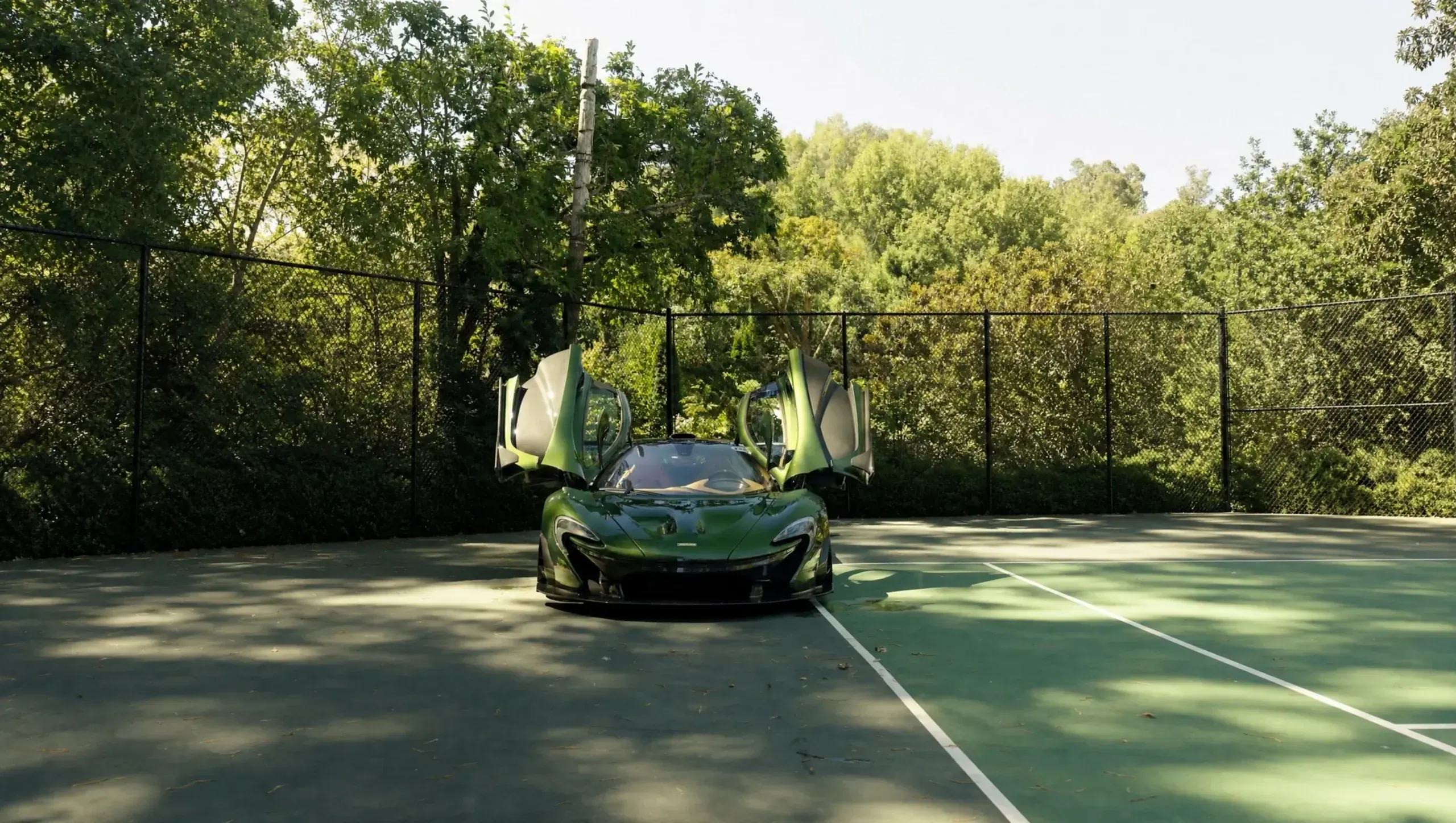 A green McLaren P1 with its doors raised, parked on a tennis court among trees